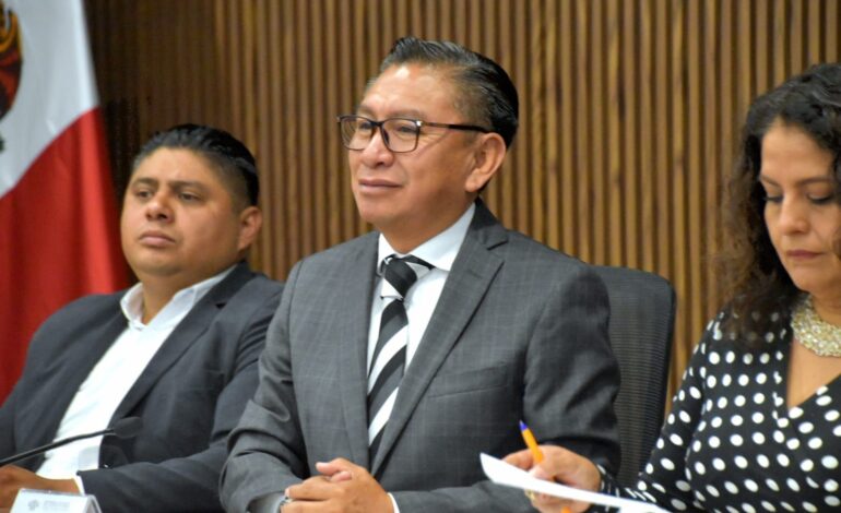 Three panelists seated at a conference table; the central man in a gray suit and glasses, flanked by a man and a woman taking notes. A flag and wood-paneled wall behind them.
