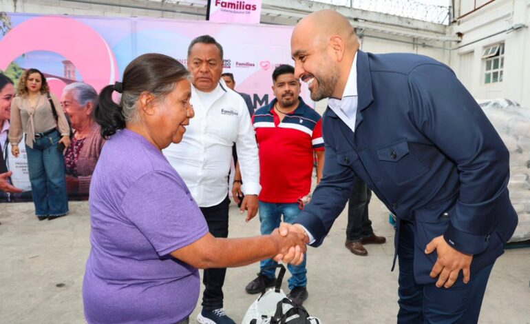 Man in a dark blue suit shakes hands with a smiling woman in a purple shirt at a community event, with onlookers and banners in the background.