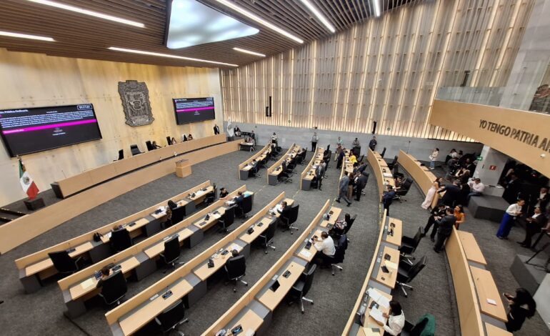 Wide view of a government chamber with curved desks, attendees seated, and large screens at the front wall
